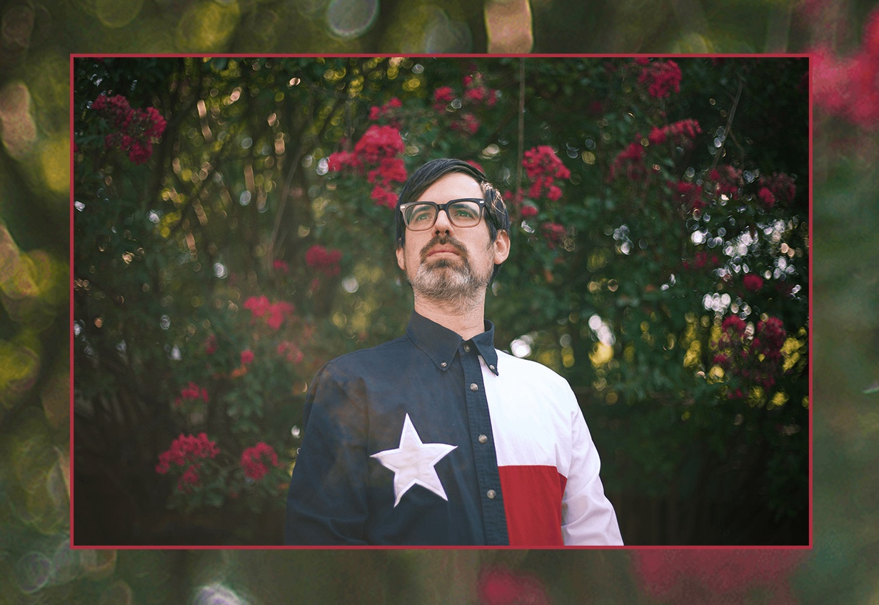 A man with glasses in front of a rose bush. He wears a shirt with the Texas flag on