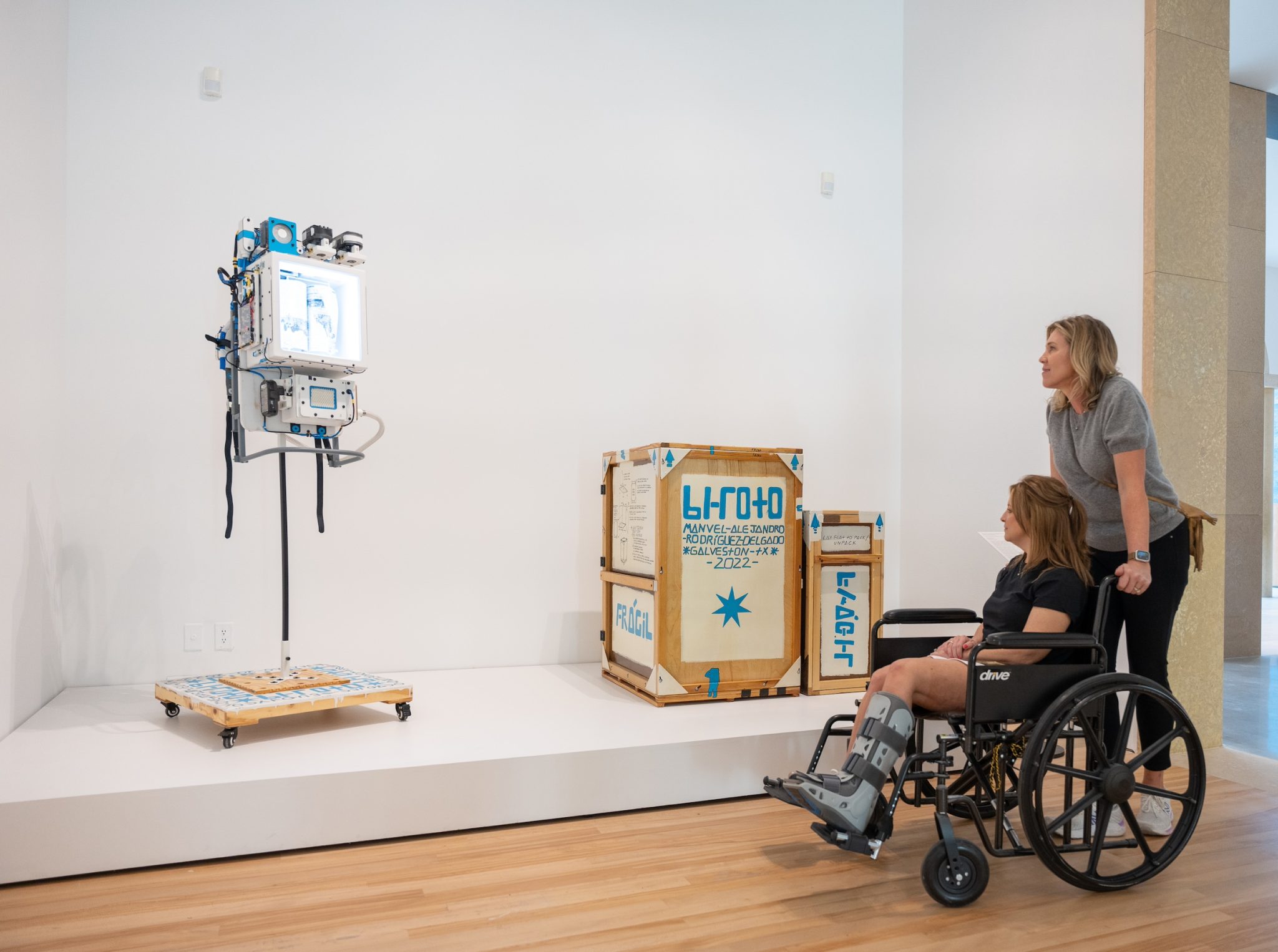 A person pushing someone wearing a leg brace in a wheelchair, looking at art in a gallery space