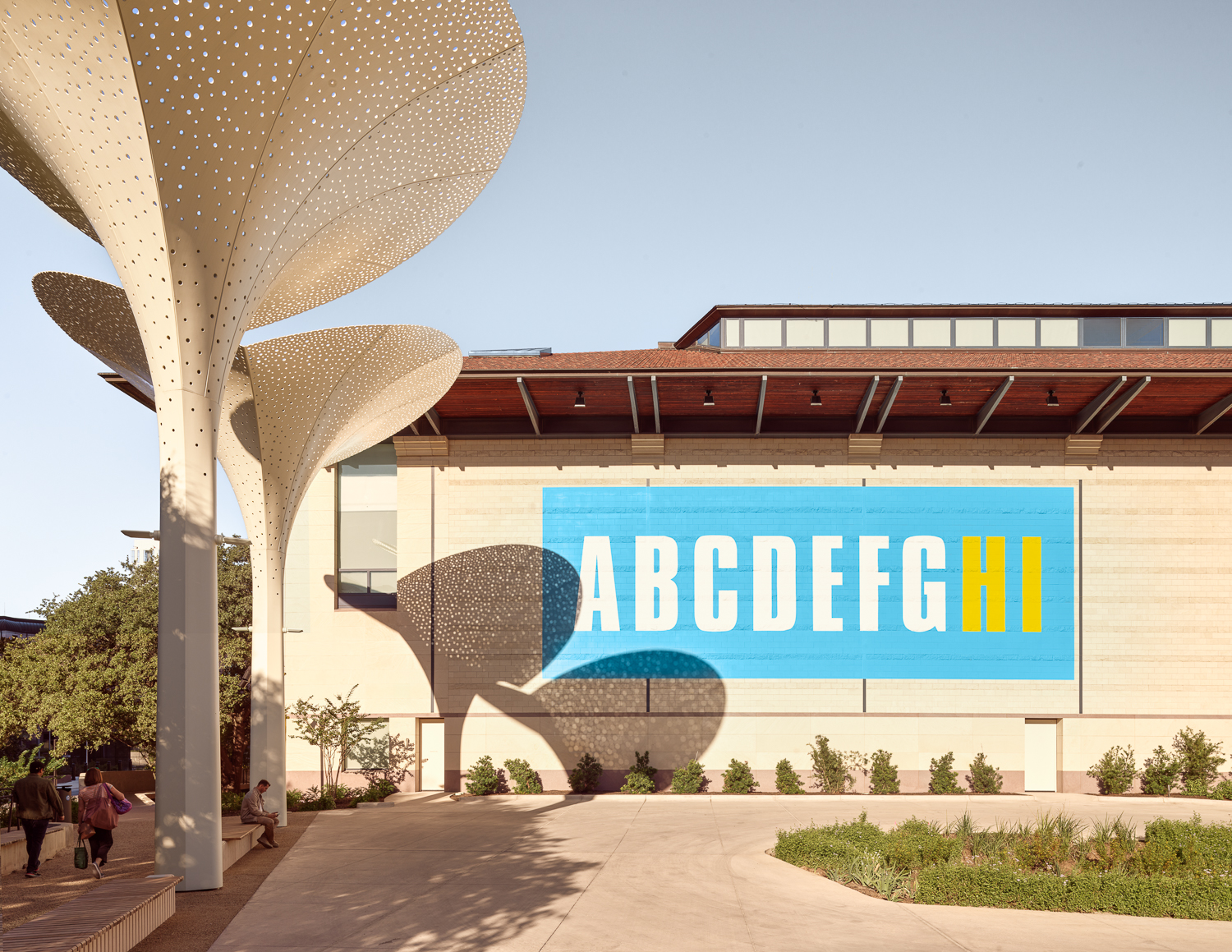 Exterior view of a modern building with a large blue banner featuring the alphabet letters "ABCDEFGHI" prominently displayed, with the 
"HI" in yellow, contrasting the white of the other letters. The architecture includes unique, canopy-like structures creating patterned shadows on the ground.
