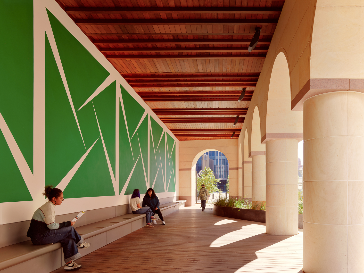 Individuals seated and walking under a covered walkway with arches, featuring large green geometric shapes on the wall, outside the Blanton Museum of Art