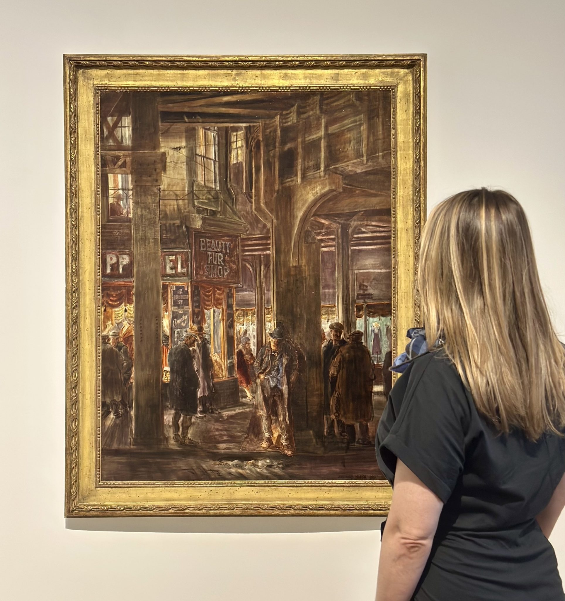 A person with long blonde hair looking at a framed artwork featuring a painting of a busy New York shopping square set in the 1930s