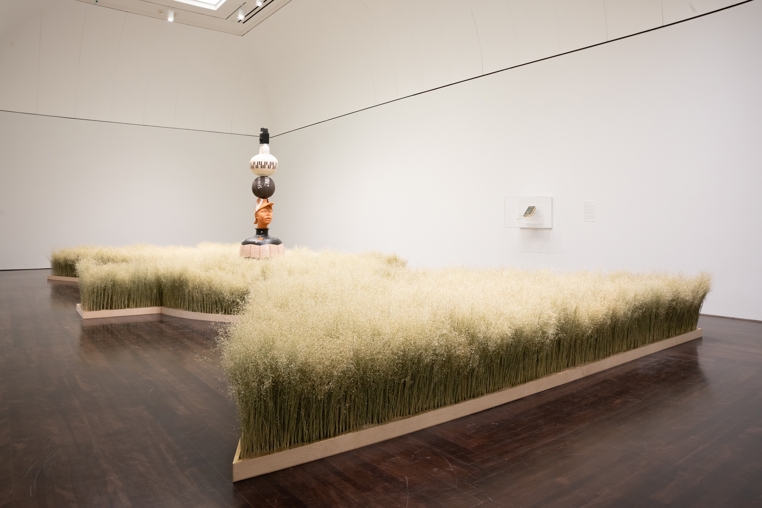 A large sculptural art installation, containing dried rice grass that fills a lot of the room. At the center is a tall clay sculptural work containing a vase at the base, topped with a bust of Marcus Garvey, a basketball, a kettle bell shape and a camera