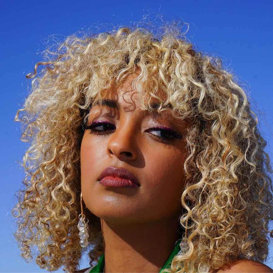 A black woman with curly blond hair and a blue sky background