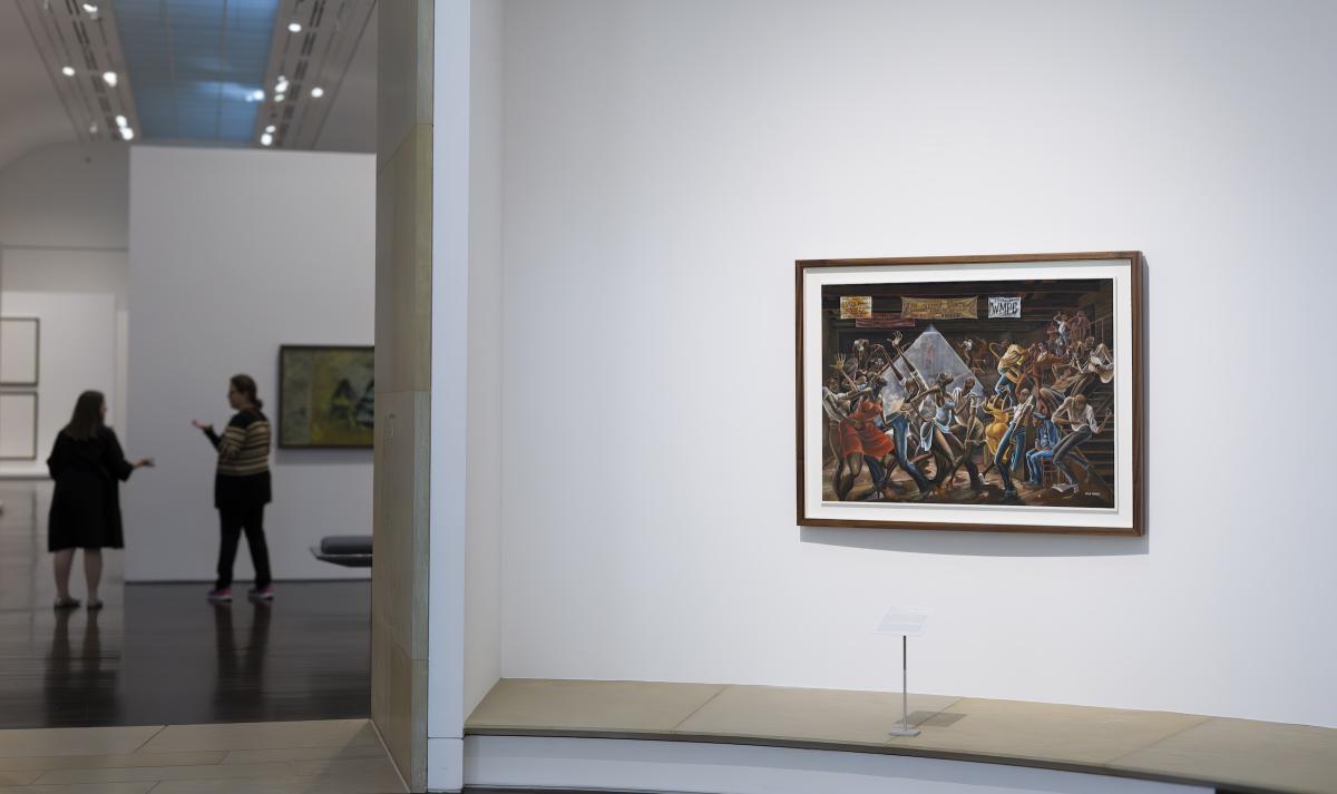 A framed painting on a gallery wall. In the background are two people standing and talking