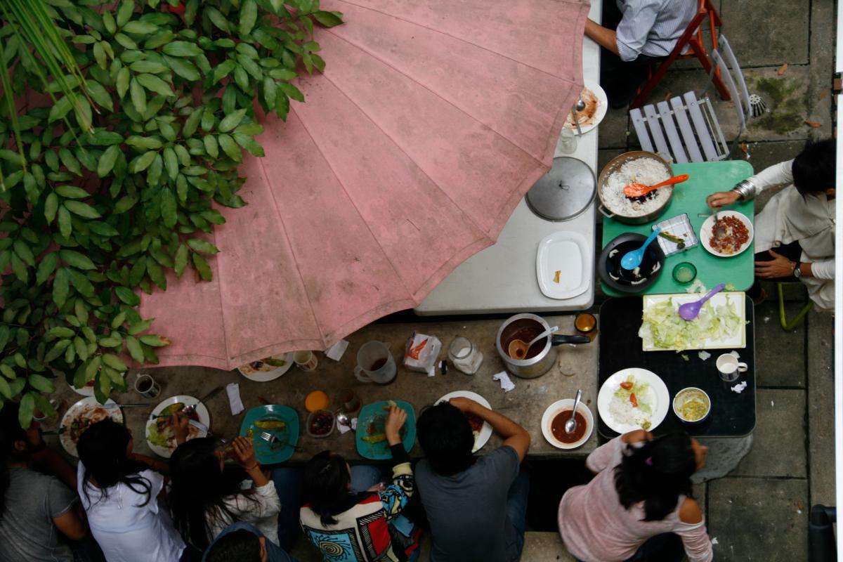 Fotografía tomada desde arriba, de una mesa con comida y personas sentadas a su alrededor. En primer plano, hay hojas de un árbol y una sombrilla rosa.