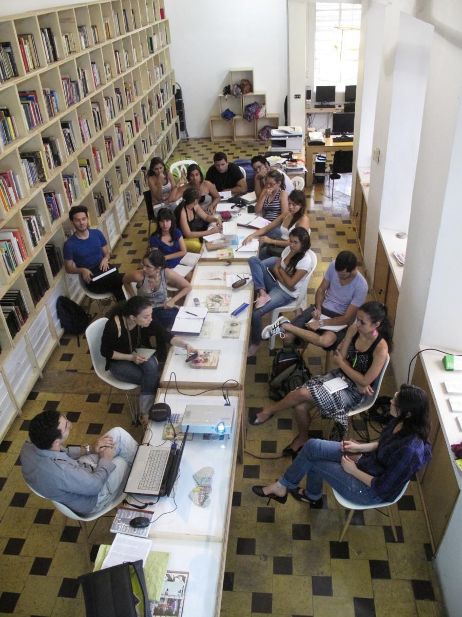 A long, white table with people seated around it. On the left side, the room has bookshelves.