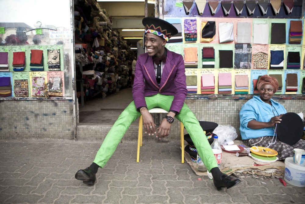 Lethabo Tsatsinyane wearing a purple jacket and green pants sitting on a stool photographed for Dazed magazine, from the Smarteez series