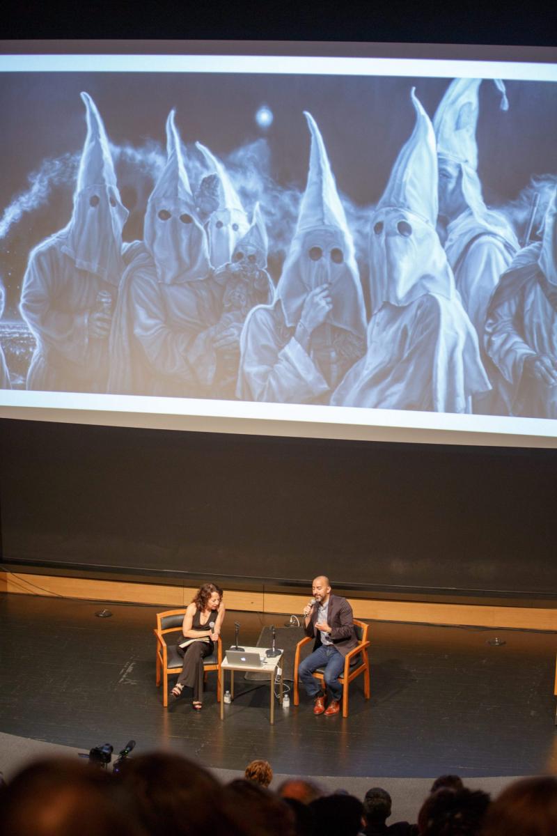 Vincent Valdez discussing his painting, "The City," with Maria Hinojosa in an auditorium. A large close up of the painting is projected on a screen behind them.