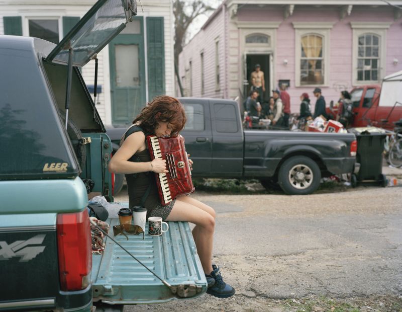 Woman sitting on a truck tailgate with two coffee cups and one mug. She is playing an accordion. In the background there are two shotgun houses and a group of men.
