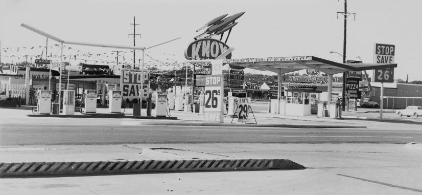 Black and white photograph depicting a roadside gas station. There are several 60s-era gas pumps on a concrete island, and four signs saying various combinations of "Stop," "Save", and "26.9." There is a large oval sign in the middle of the photograph with the word "Knox" shown in capital letters. Mounted at the top of the sign is a 50s depiction of a rocketship, with three bullet-shaped rockets.