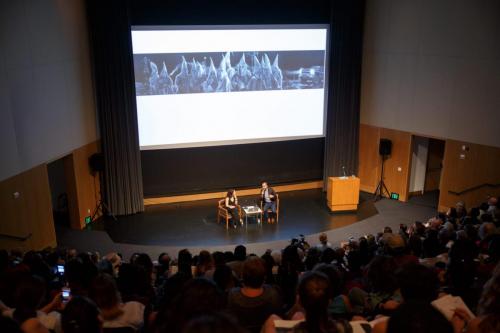 Full auditorium view of Vincent Valdez discussing his painting, "The City," with Maria Hinojosa. An image of the painting is projected on the screen behind them.