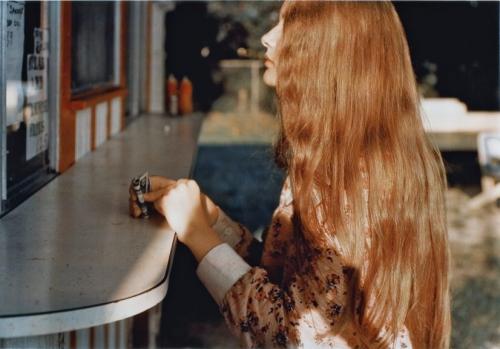 A younger woman with long red hair is in the foreground, facing to the left. She is holding money in her right hand, and both of her hands are on a counter. She appears to be standing outside at a window preparing to order food.