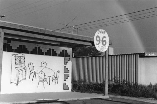 A concrete overhang with a prominent circular sign advertising "Open 9 to 6, Closed Sun" is attached to the right side of the overhang. Under the overhang is a large drawing of a table and chairs set, as well as a china hutch. The drawing is surrounded by graphic blocks. In the background, a rainbow is visible through several power lines.