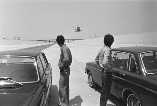Black-and-white photograph depicting two cars, two men facing away from the camera, and a helicopter in the distance framed between the two men. A shallow river is visible on the left. The remainder of the landscape is concrete. A bridge is visible in the distance.