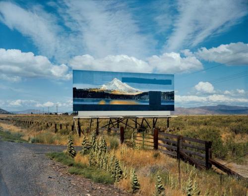 A billboard depicts a snow covered mountain, lake, and pine tree forest. A barbwire and wooden fence separates the billboard from the road. The land is flat and grassy with hills in the background.