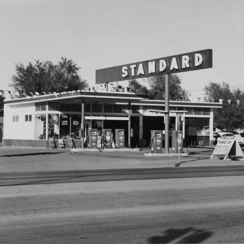 A road crosses in front of the camera in the foreground. Across the road is a gas station, with a large sign with the word "STANDARD" mounted above it. There are two concrete islands, one with two gas pumps and one with three. There is a garage attached to the station, with three open bays. Signs for tires and tune-ups litter the area.