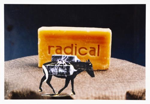 The piece is made out of a collage of photos, paper and stamps. An orange soap bar with radical carved into it sits on a brown cloth covered table. In front of the soap bar is pasted a donkey with cargo strapped on its back.