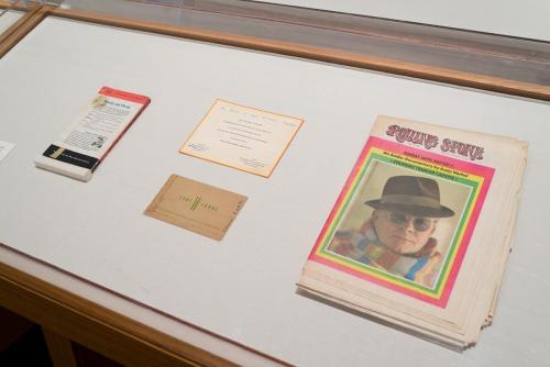 Back cover of a book, a copy of Rolling Stone magazine and two printed cards with some handwritten notes in a display case
