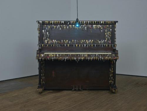 An upright piano decorated with keys. A hanging blue bulb is positioned in front of it.
