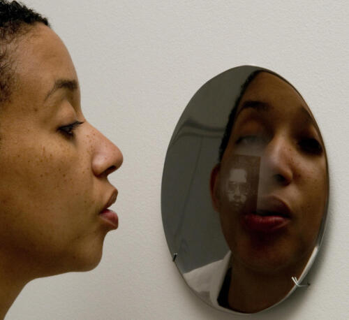 Color photograph of a person blowing on a small round mirror. The mirror shows two images: the reflection of the face of the person blowing on the mirror, and a small square with a black and white portrait.