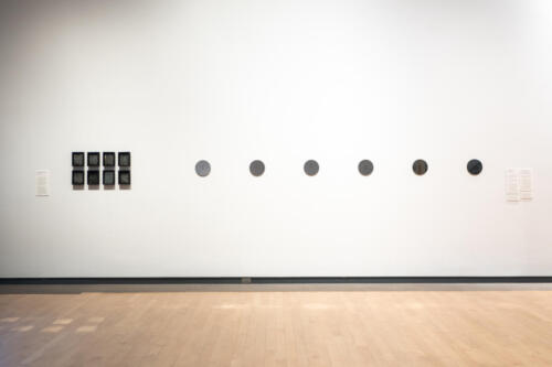 A white wall and wood floor. On the left of the wall, there are eight small black frames in two horizontal lines of four frames each. On the right side, there are six small round mirrors forming a horizontal line.
