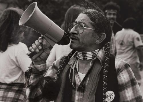 A man with plaits wearing glasses and a plaid shirt, speaks into a megaphone at a rally. His expression is focused and determined, his mouth open as he speaks. Other attendees can be seen in the background, blurred and indistinct.