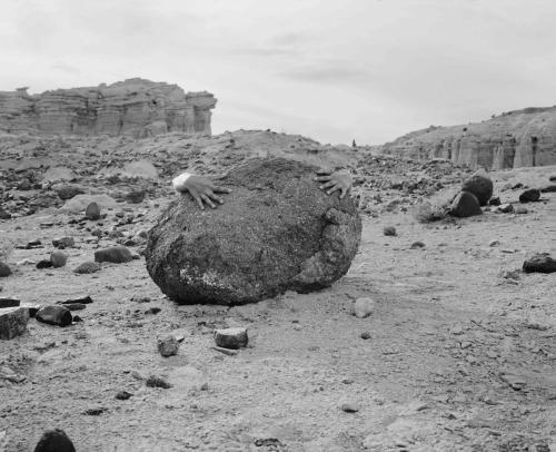 A black-and-white photograph of two hands embracing a large rock in an otherwise empty desert