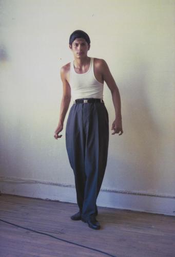 A person in a white tank top and high-waisted dark trousers stands confidently in a sparsely decorated room with wooden floors.