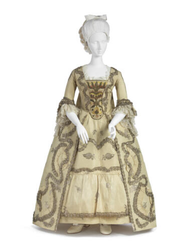 European beige dress on a mannequin. It is decorated in rich embroidery at the chest, with lots of lace at the low neck and fluffy sleeves. It has an opening on the skirt that shows the undergarments made in the samefabric.