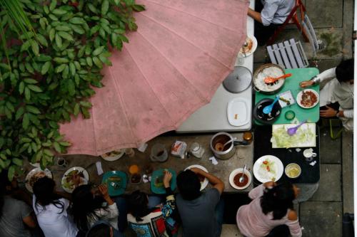 Photograph taken from above, of a table with food and people seated around it. On the forefront, there are leaves of a tree and a pink umbrella.