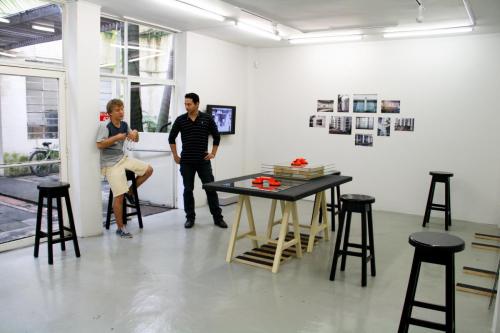 Two people inside a white, light room with tables and chairs. On the wall, there are images hanging.