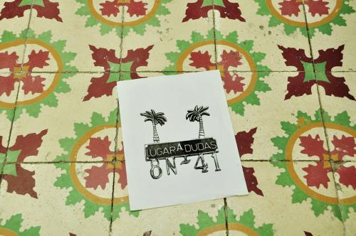 A rectangular piece of white paper on a colorful floor. The paper has two palm trees, and it says: “Lugar a dudas. 8N 41”